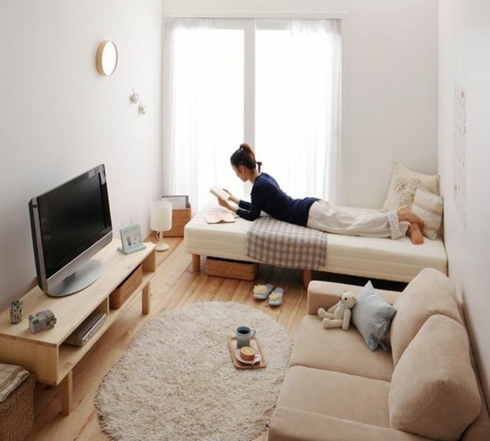 brunette young woman, lying on a narrow white bed, inside a small room with white walls, and a window, couches for small living rooms, beige sofa opposite a tv, on a wooden stand 