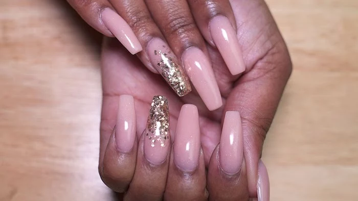 ballerina nail shape, painted in nude pink, and decorated with flakes of golden glitter, on brown hands, resting on a light wooden surface