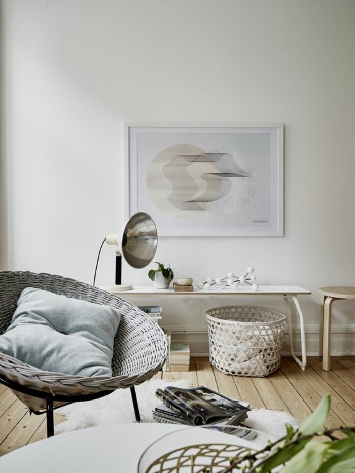 abstract painting mounted on a white wall, inside a room with pale, beige wooden floor, containing an off-white wicker chair, a white desk and a stool