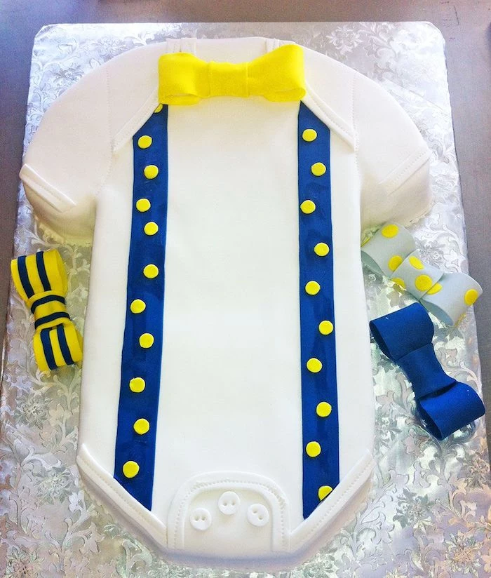 bowties in yellow and dark blue, plain and patterned, decorating a onesie cake in white, with blue suspenders, and three white buttons