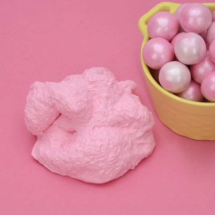 wad of lumpy, bubblegum pink fluffy slime, placed on a pink surface, near a yellow plastic container, filled with pearl-like pink marbles