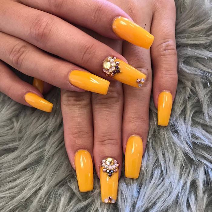 burnt yellow nail polish, decorated with rhinestones, on the long coffin nails, of two hands, resting on a fur-like, pale grey surface