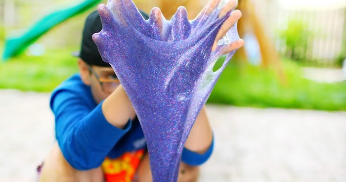 boy playing with purple, glittery slime without borax, stretching it between his hands, blue and red top, baseball cap and glasses