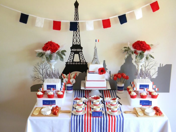 flags in black white and red, hung above a table with a white red and blue tablecloth, containing various French sweets, 2 bouquets of red roses, a three layered cake, 60th birthday ideas, two cheese boards, and an eiffel tower mural