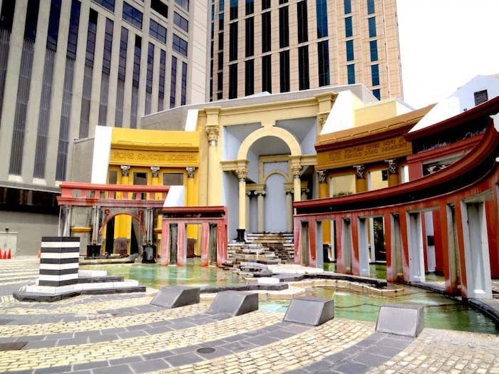 roman theatre-resembling structure, with elements painted in white, red and yellow, and a shallow pool, skyscrapers in the background
