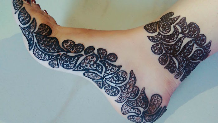 dark henna foot tattoo, on the side of a foot, and around the ankle, asymmetrical outlines filled with small flourishes