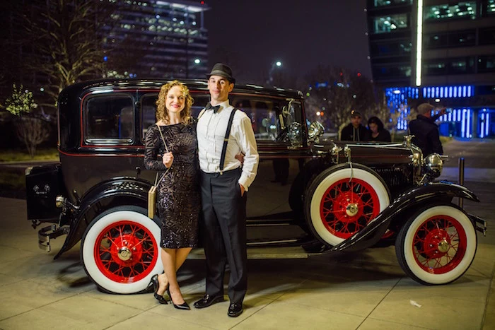 curly blonde woman, wearing a 1920s, art deco-inspired black dress, standing next to a man, in black trousers and white shirt, with suspenders and a bowtie, retro car in the background, great gatsby mens clothing