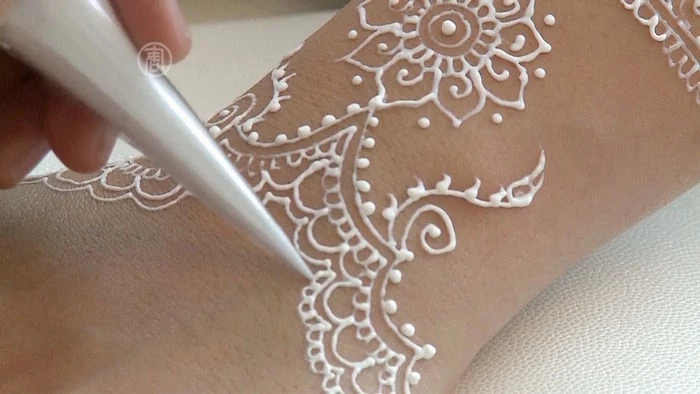 silver cone held by a hand, applying white artificial henna, on a pale wrist, henna tattoo with floral motifs, and symmetrical details