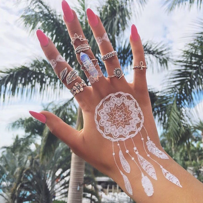 dreamcatcher-shaped, small henna tattoo, in pure white, on a hand with many different boho rings, and sharp pink nails