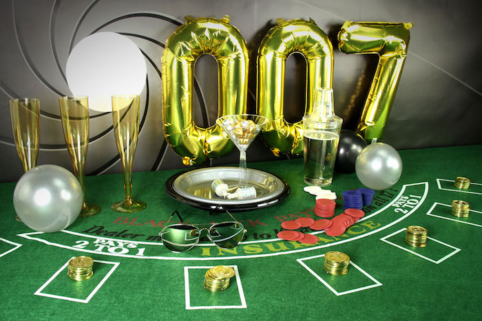 gambling table with chips, gold coins and three yellow champagne flutes, metallic balloons shaped like the number 007, 60th birthday party ideas for men, james bond theme