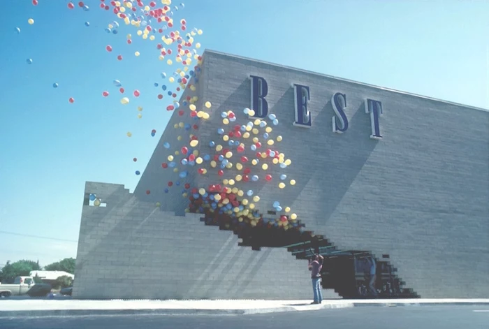 pale grey brick building, featuring the word best, written in large blue letters, on one of its walls, many balloons in different colors, flying out of a large crack in the building