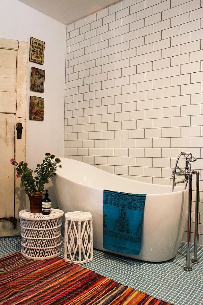 bathroom decorating ideas on a budget, white subway tiles on the wall of a room, containing a white modern bathtub, pale blue mosaic floor, and a multicolored striped rug, flowers and white rattan furniture