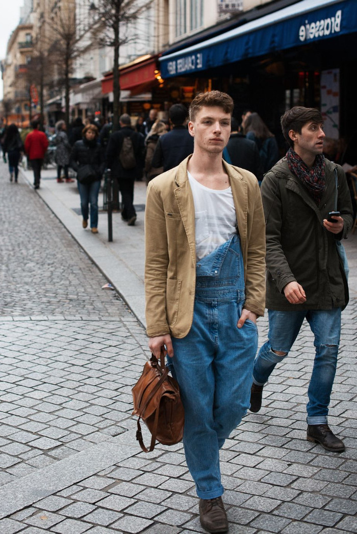 blazer in beige, with long baggy, half unbuttoned denim overalls, worn by a young man, holding a brown leather bag
