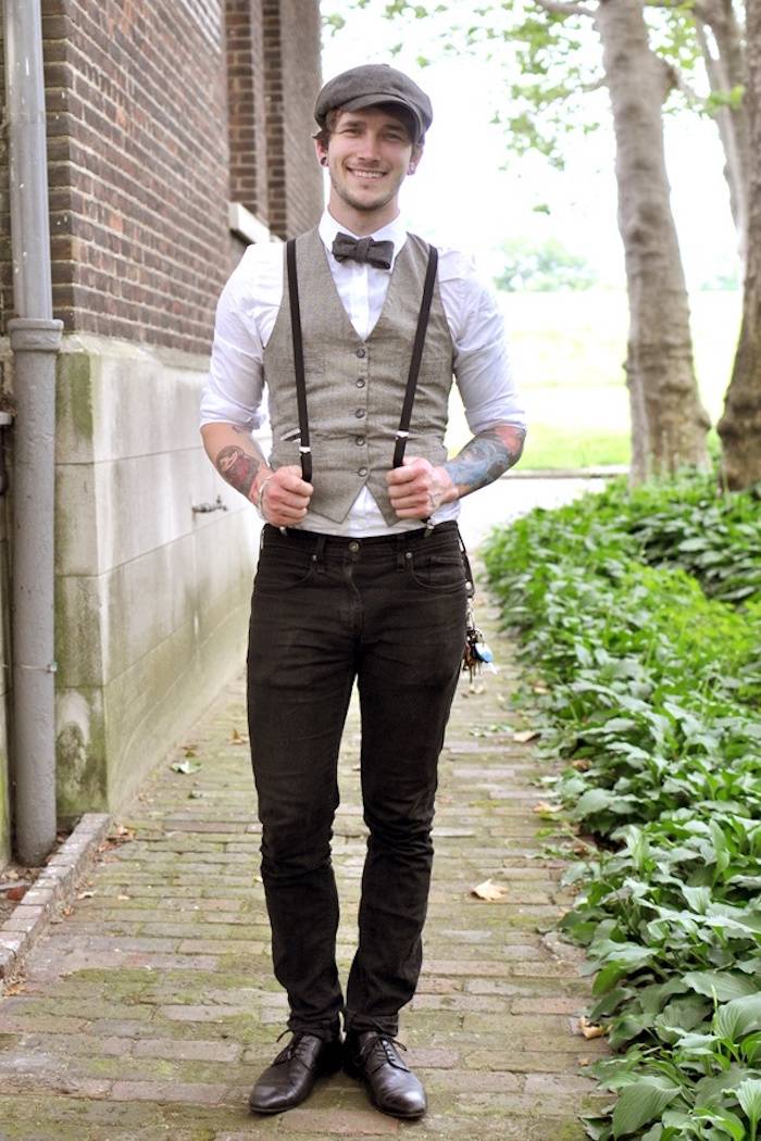 trousers in black, worn with suspenders, a white shirt and a beige vest, and a brown bowtie, 20s mens fashion, on a smiling man with a cap, and forearm tattoos