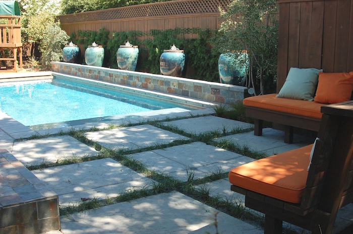 five large ornate vases, decorating a small wall, near a light blue pool, small inground swimming pools, garden with two wooden benches, covered in orange and pale blue cushions