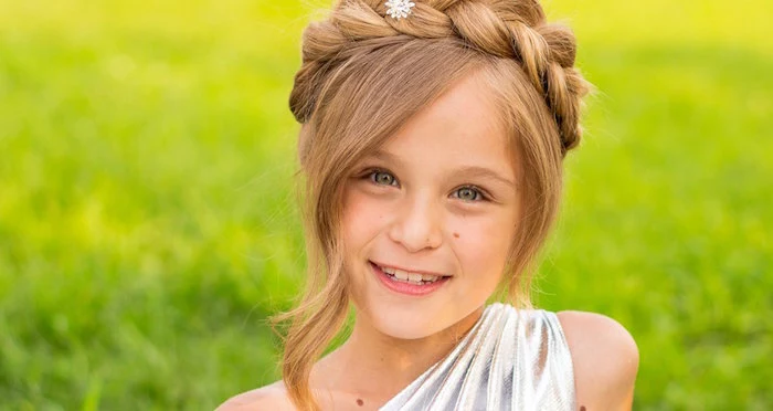 honey blonde child, with long curled side bangs, and a crown braid, cute girls hairstyles, smiling in a silver, one-shoulder dress