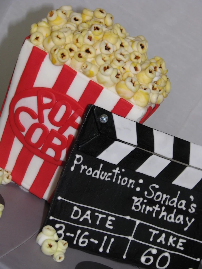cake shaped like a vintage popcorn box, in red and white, behind a black clapper board, both made from colorful fondant, 60th birthday party ideas, film-themed cakes
