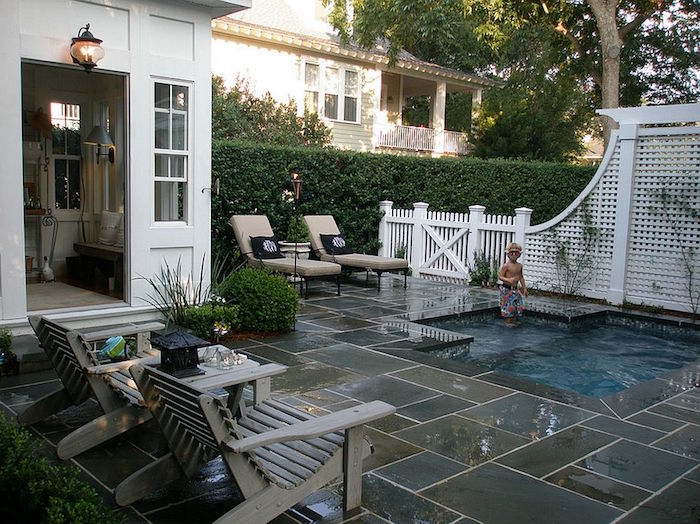 house with an open door, near a yard, covered in grey and beige stone tiles, containing a pool, with a child playing, cool backyards, two wooden chairs, and two sun beds