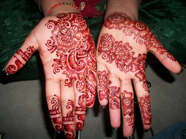woman showing her palms to the camera, they covered in red mehndi, henna hand tattoo designs, with intricate floral motifs