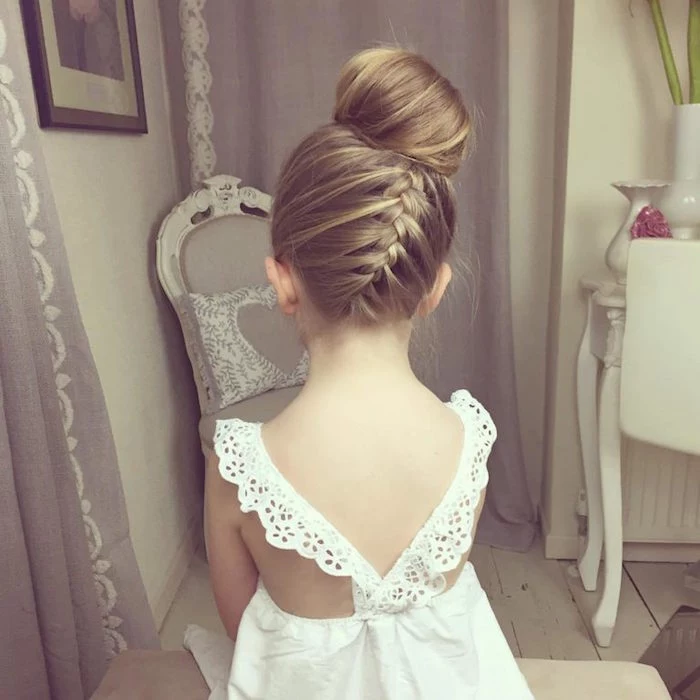 back view of a braided hairstyle, with a blonde side braid, and a bun on top of the head, worn by a small child, in a white summer dress
