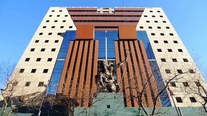 woman holding a trident, statue made of bronze, at the entrance of a tall, cube-shaped building, with small square windows, glass panels and striped details, postmodernism examples 