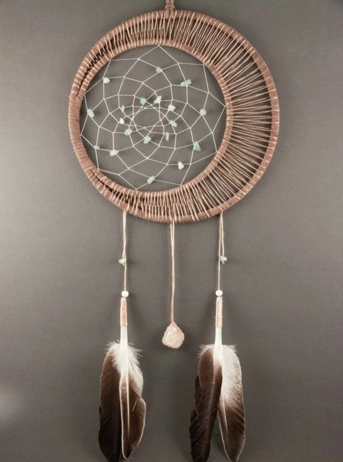 two chocolate brown and white feathers, and a small pink stone, tied to a rattan dreamcatcher, with a white net, decorated with pale blue beads, pictures of dream catchers, on a grey wall
