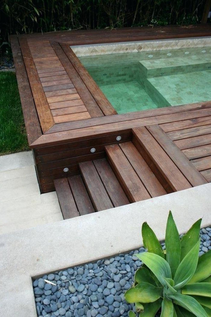 four wooden steps, leading up to a pool, surrounded by a wooden surface, pool patio ideas, flower bed with pebbles, and a green plant nearby
