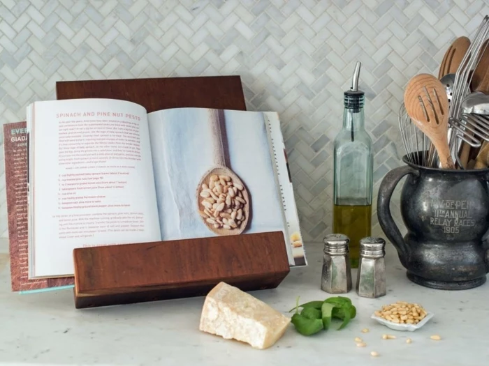 stand for cooking books, made of wood, holding an opened book, piece of cheese, basil leaves and nuts, condiments and utensils nearby