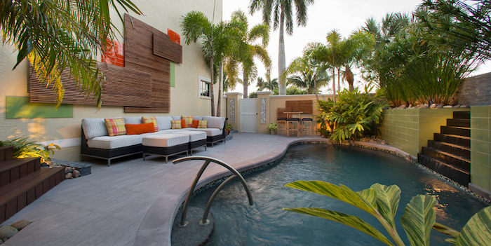 backyards with pools, black and white sofa, with two matching tables, and colorful yellow and orange cushions, in a garden with palms, other plants and a pool