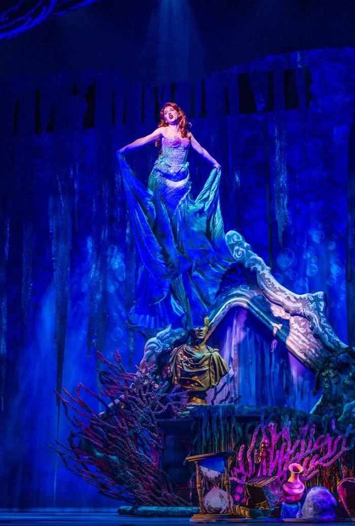 woman in a long blue gown, and red wig, standing on a stage, with elaborate set design, made to look like an underwater world