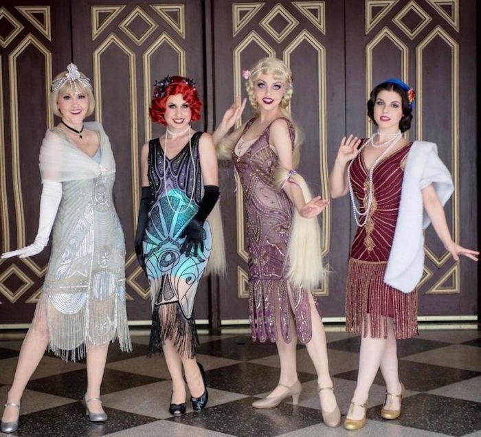 group of women, each in a different gatsby themed dress, pale blue and silver, turquoise and black, pink and purple, red and gold, all with embroidery and fringe details