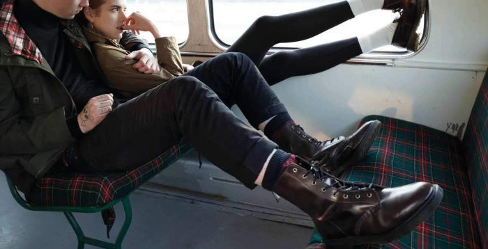 hugging young couple sitting in a bus, the boy is wearing skinny trousers, green parka and brown lace up combat boots, 90s outfit ideas, the girl is wearing black leggings, whites socks and a khaki jacket