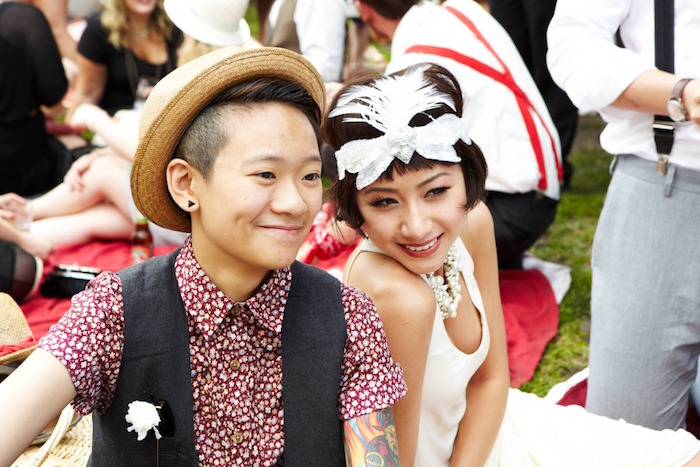 happy-looking couple, sitting next to each other, wearing a short-sleeved patterned shirt, and a black vest, and a white retro gown, straw hat and feathered headband