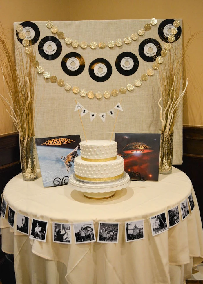 dried plants arranged in two sparkling vases, placed on either side of a white cake, with gold details, vinyl records and two boston albums, 60th birthday color, on a table decorated with black and white photos