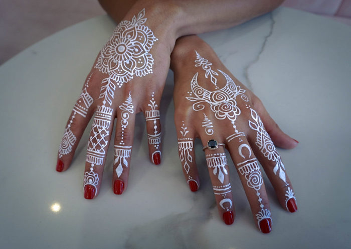 marble-like surface in white, and pale grey, under two hands, with red nail polish, decorated with white, temporary henna tattoos, with flowers and crescent moons