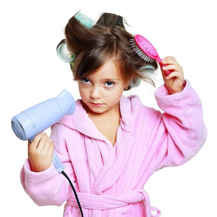 child in a pink bathrobe, with big curlers on her head, cute hairstyles, brushing her hair, with a small pink brush, while holding a blue hairdryer 