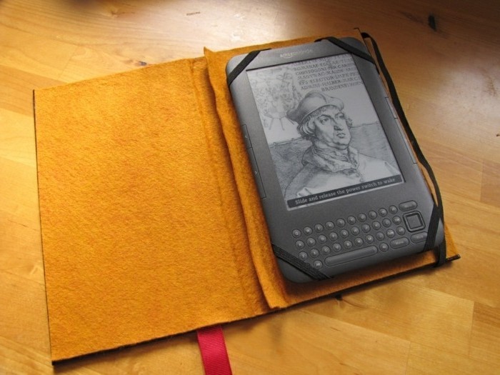 kindle inside a protector, shaped like a book, with hard beige covers, and a red bookmark, handmade gifts, placed on a laminate floor
