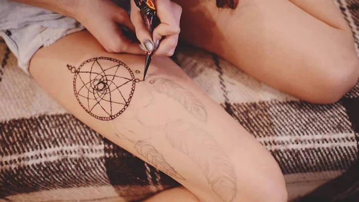 sitting slender woman, dressed in pale denim shorts, drawing a dreamcatcher on her thigh, using a cone of henna, cute henna designs