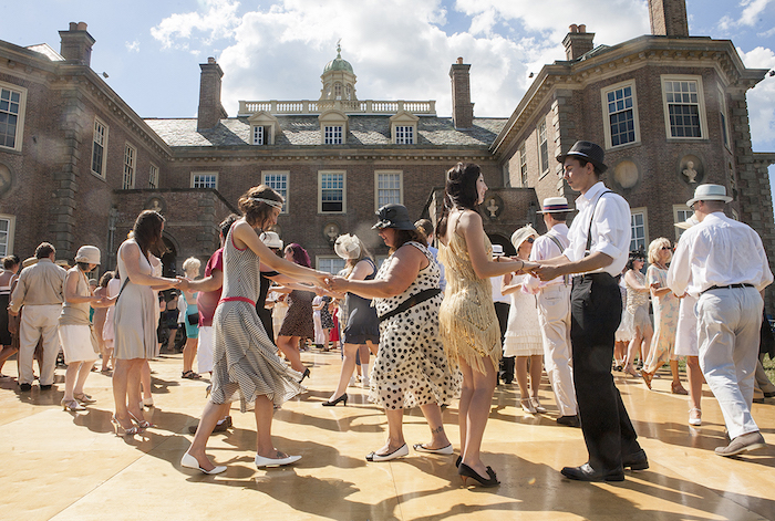 brick mansion, standing behind a croup of dancing people, all dressed in roaring 20s fashion, flapper dresses and headbands, trousers with suspenders and shirts