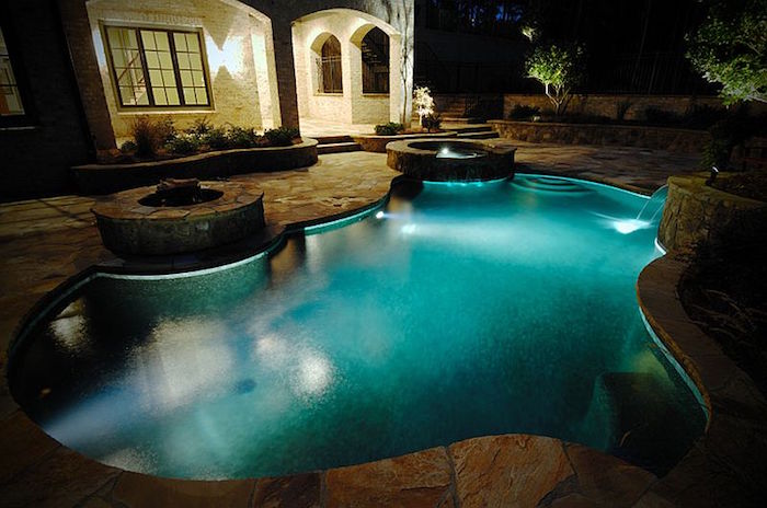night photo of a pool, lit from within, near a fire pit, in the back yard of a house, with a lit porch light