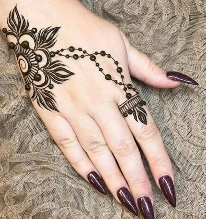 beige lace under an arm, with long sharp nails, painted in a dark plum nail polish, a black henna tattoo, featuring a flower and a ring-detail