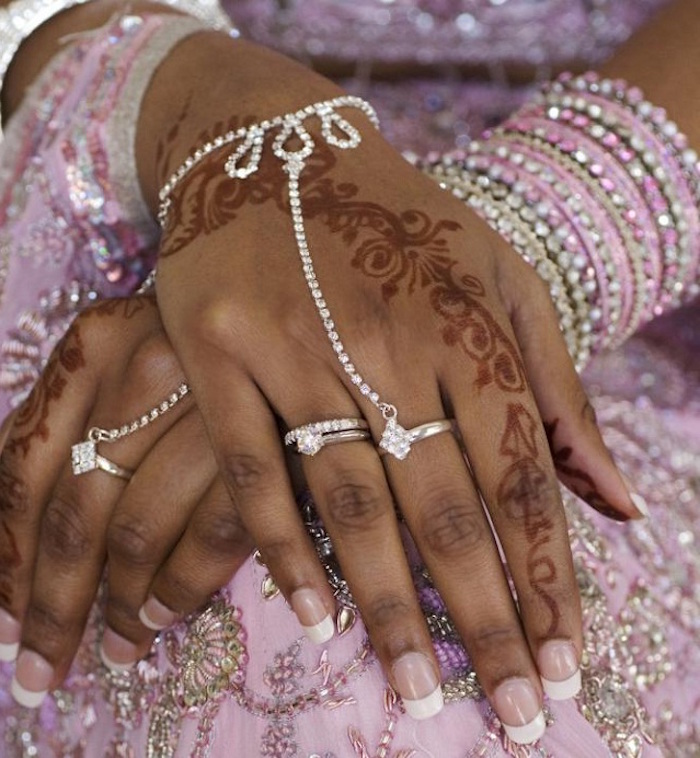 brown hands decorated with rings and bracelets, a french manicure, and long finger henna tattoos, resting on shiny, pale pink embroidered fabric 