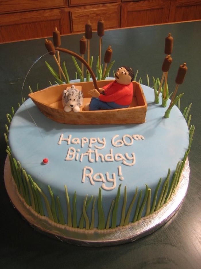 man with a fishing rod, and a small dog in a boat, decoration made from colourful fondant, on top of a pale blue cake, with grass and fondant reads, happy 60th birthday ray, written in white