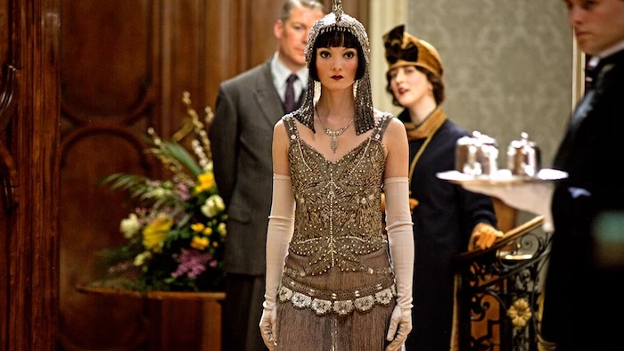 headdress in shimmering silver, on a woman with short black hair, and bold make up, wearing a beige and gold flapper gatsby dress, with pearls and beads