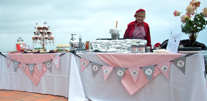 woman dressed in red, standing near two tables, with white and pale pink tablecloths, 60th birthday party ideas for mom, cupcakes and a crepe station