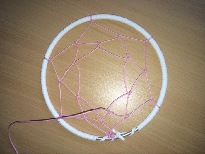 web-like pattern, made with pink thread, inside a white hoop, how to weave a dreamcatcher, placed on a light wooden surface