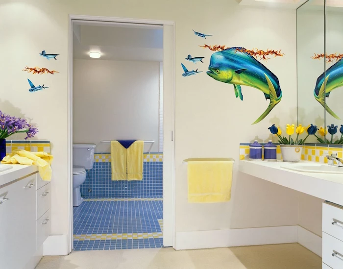 one big and several small fish decal stickers, on the pale yellow wall, of a room containing white cupboards, floral decorations and yellow towels, modern bathroom ideas, open door revealing a toilet in the next room