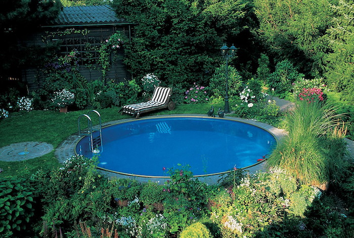 trees and shrubs, flowers and grass, in a green garden, containing a blue circular pool, striped sun bed nearby