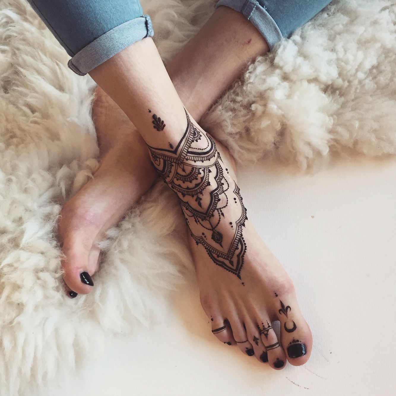 sheepskin rug in creamy white, under a pair of feet, one of them decorated with an intricate, black henna tattoo, black nail polish 
