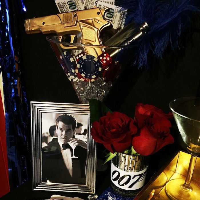 photo of piers brosnan, holding a martini glass, in a silver frame, next to a large decorative glass, containing a toy gun in gold, gambling chips and dices, and faux money, 60th birthday party ideas for men, 007 themed celebration